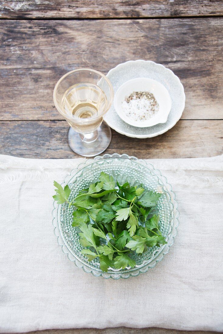 Parsley in a glass bowl with a glass of wine and a salt and pepper mixture behind it