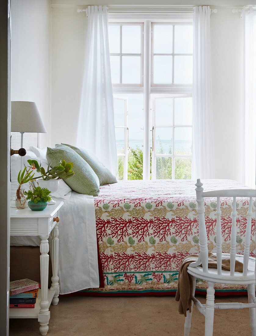 View through open door of double bed at comfortable height with floral bedspread, lattice window and airy curtains