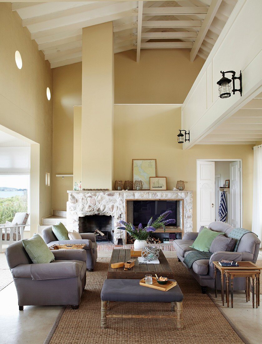 Grey sofa and armchairs around wooden coffee table in double-height interior with fireplace