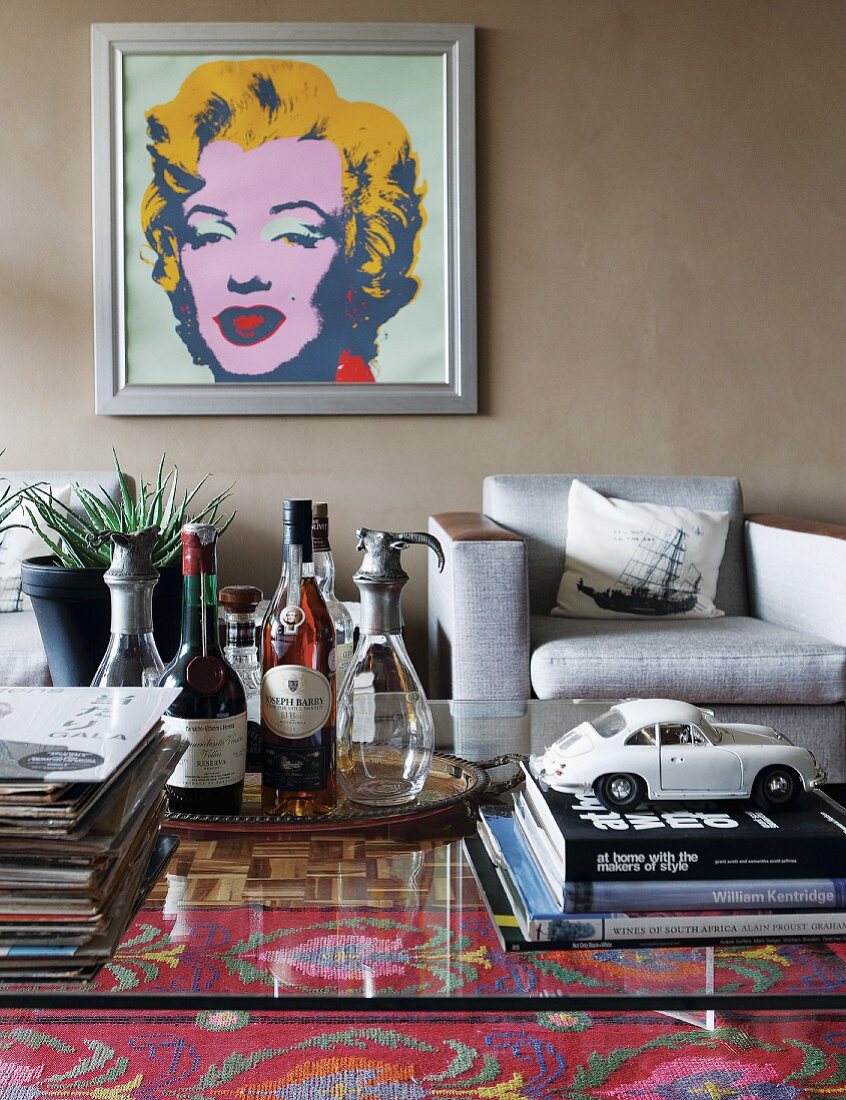 Bücherstapel und Retro-Blech-Spielzeugauto auf Glastisch, graue Sessel an schlammfarbener Wand mit Portrait von Marilyn Monroe