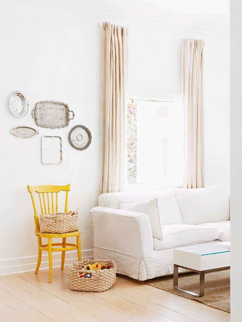 Basket on floor, yellow-painted chair and collection of silver trays on wall next to white sofa below window with floor-length curtains