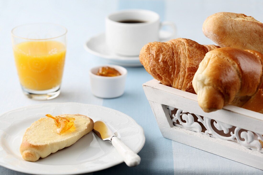 Croissant mit Butter und Orangenmarmelade, Orangensaft, Kaffeetasse, sowie Brotkorb mit Croissant, Hörnchen und Brötchen