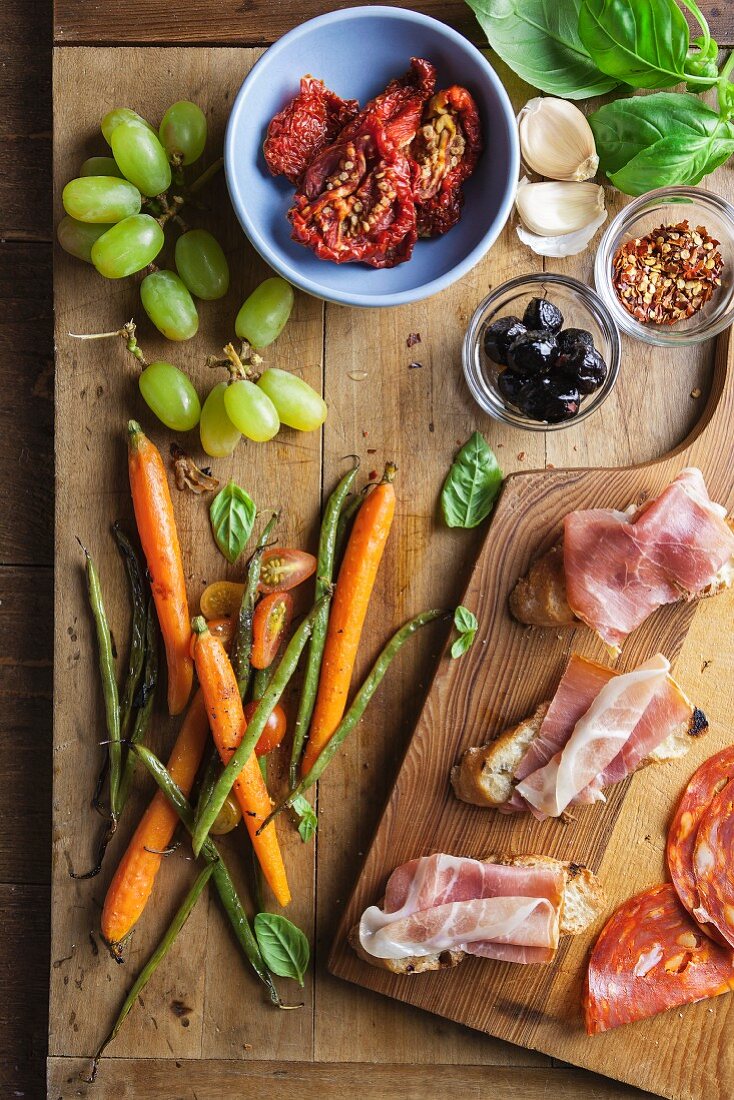 Ingredients for appetizers on a chopping board: Prosciutto, carrots, asparagus, dried tomatoes, olives, grapes basil