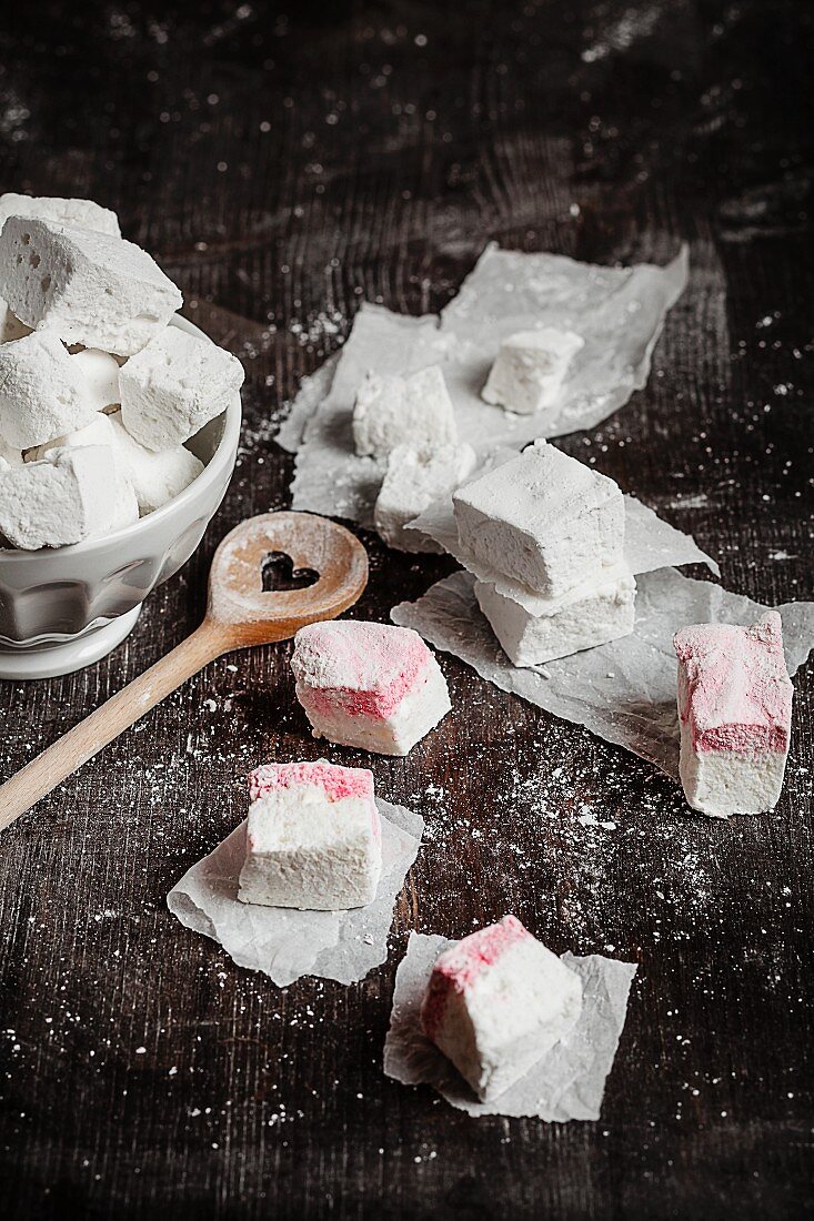Homemade marshmallows and peppermint marshmallows on a wooden surface with a wooden spoon sprinkled with icing sugar