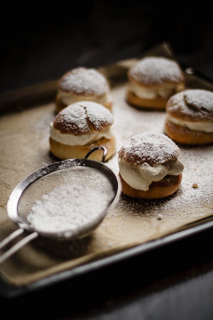 Semlor (Swedish cakes) on a baking tray next to a sieve of icing sugar