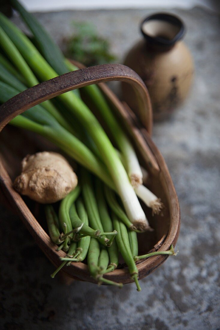 A basket of spring onions, beans and ginger next to a Japanese vase