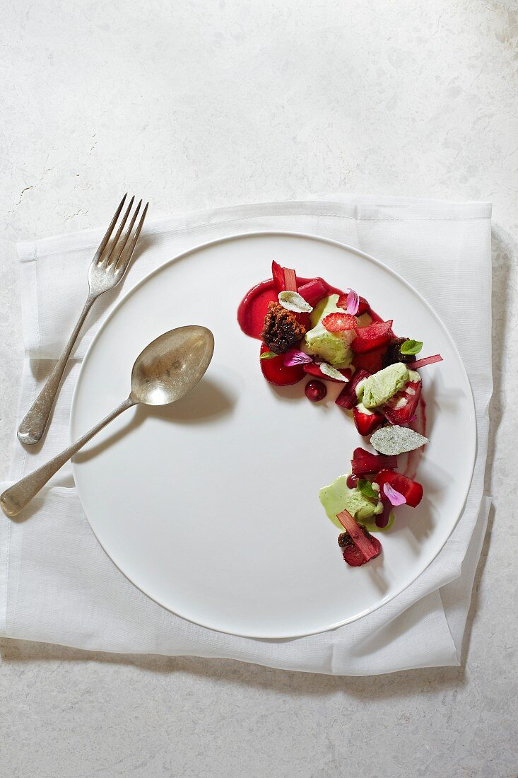 Basilikum-Semifreddo mit Siruptarte, Erdbeeren und Rhabarber auf weißem Teller und Silberbesteck