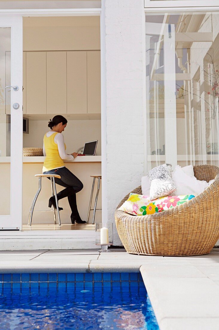 View from courtyard with pool and wicker armchair through open terrace door of woman in house interior