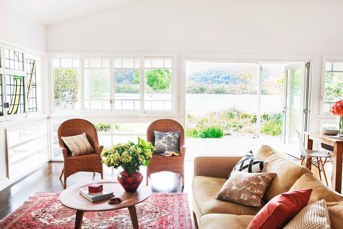 Sofa mit Kissen und Rattanstühle um Couchtisch im 50er Jahre Stil vor Fensterfront, Blick durch offene Terrassentür auf den See