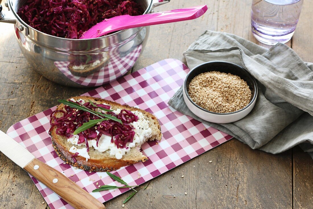 Brotscheibe mit Frischkäse, Rotkohl, Rucola und Sesam auf kariertem Frühstücksbrettchen