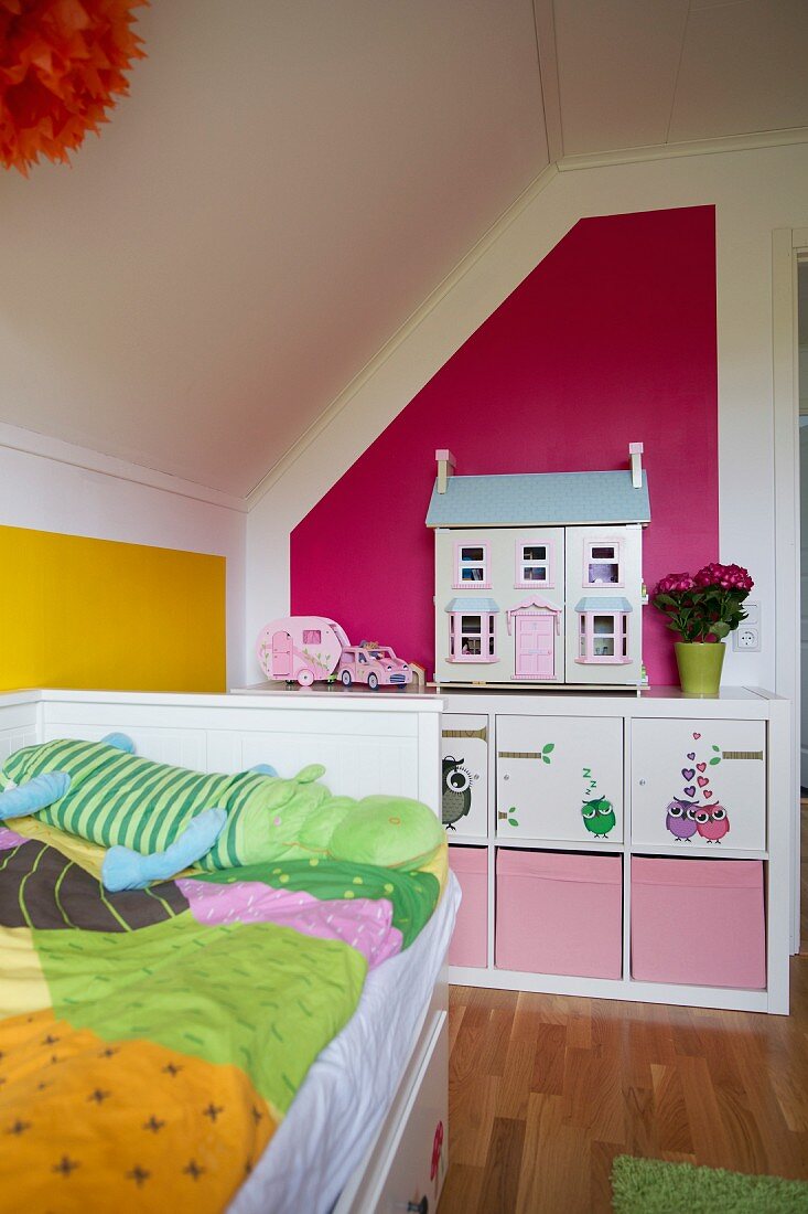 Bett mit bunt gemusterter Bettwäsche unter Dachschräge, im Hintergrund Regal mit Aufbewahrungsboxen und Puppenhaus vor pinkfarbener Wand