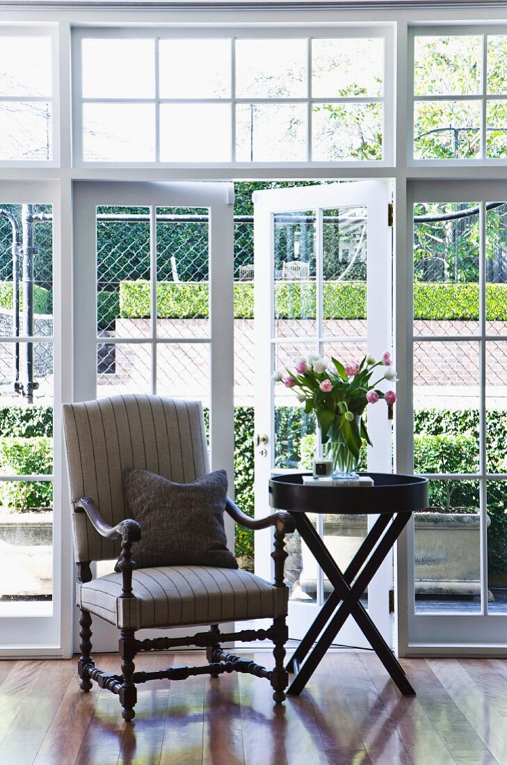 Antique armchair with turned wooden frame next to dark wooden side table in front of French windows