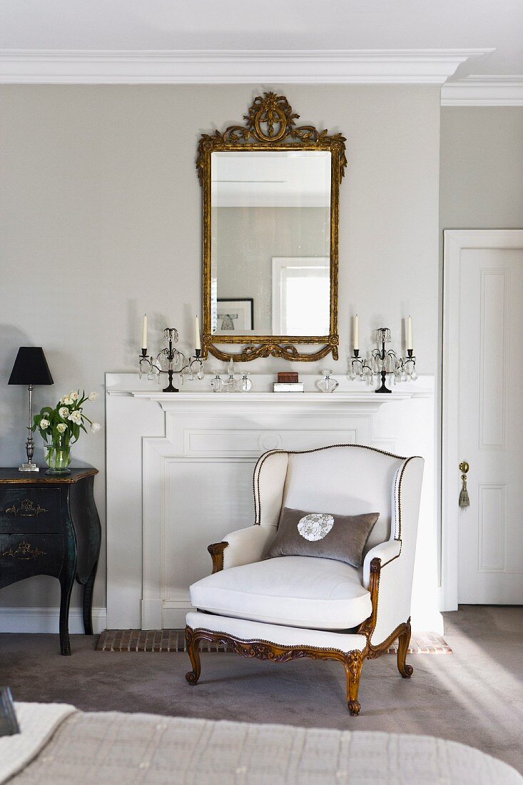 Elegant, white Rococo-style reading chair in front of fireplace with wooden surround in traditional interior