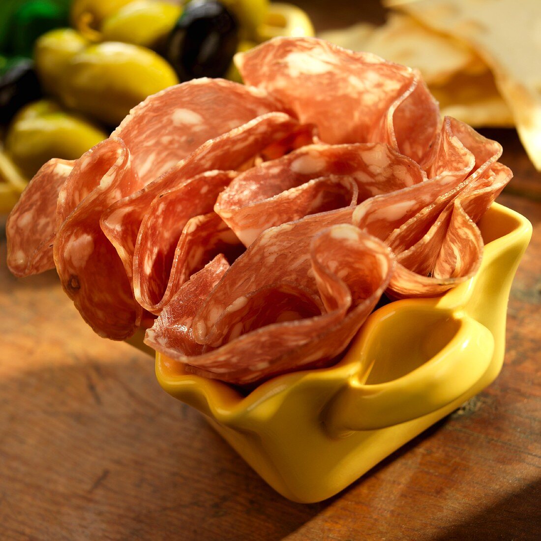 Sopressata in a yellow dish with green and black olives and crackers