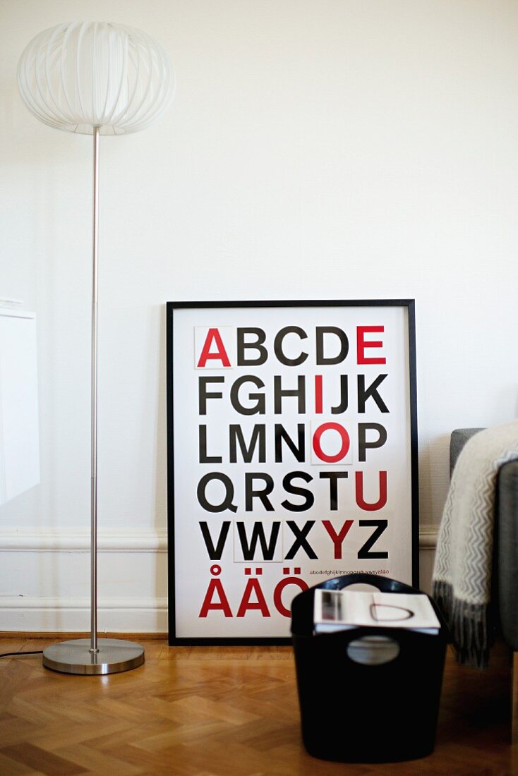 Black bag on herringbone parquet floor in front of framed alphabet next to retro standard lamp