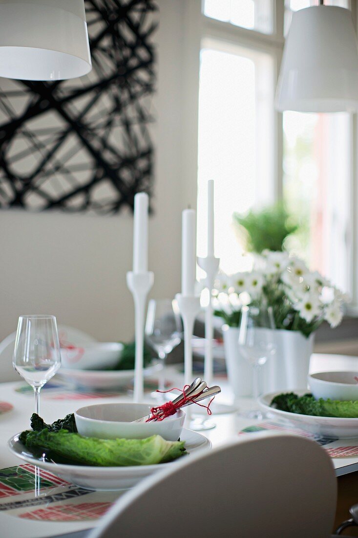 Place settings with white bowls on arrangements of cabbage leaves and white candlesticks on table