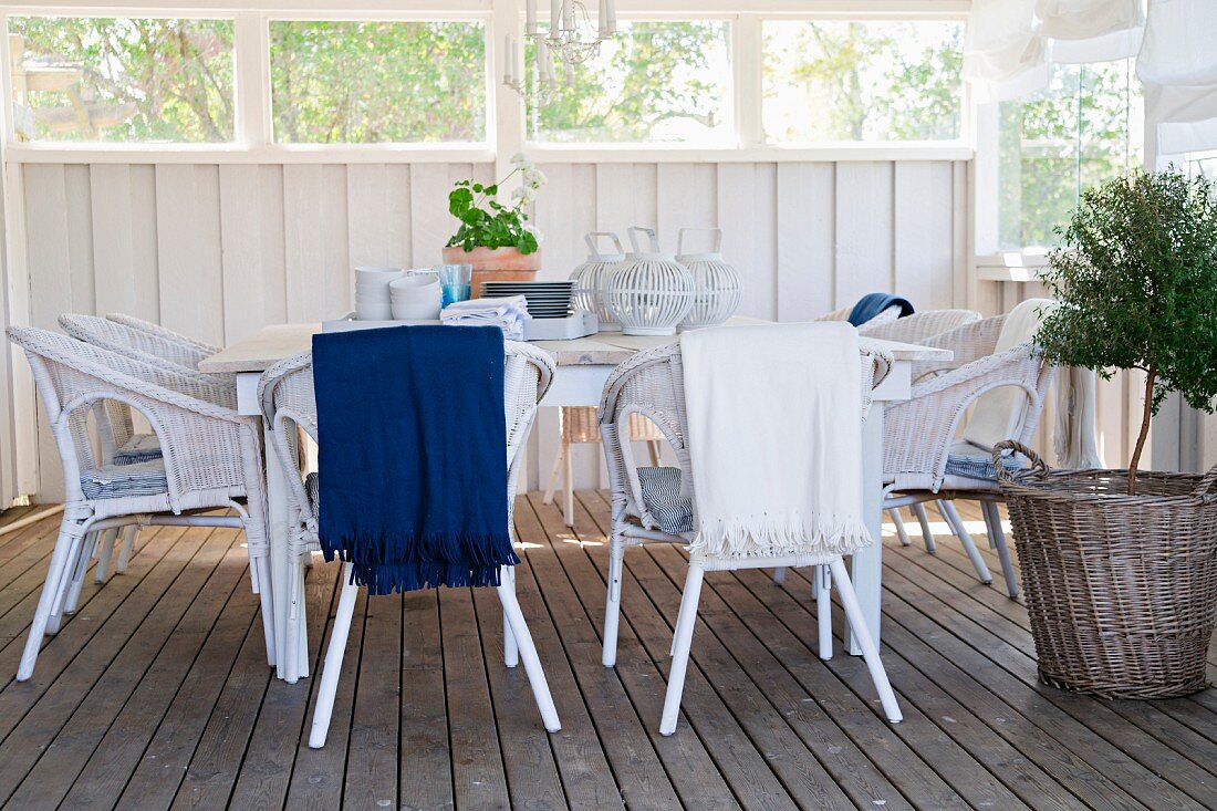 Seating area with white wicker chairs next to small tree in basket on wooden floor of wood-panelled veranda