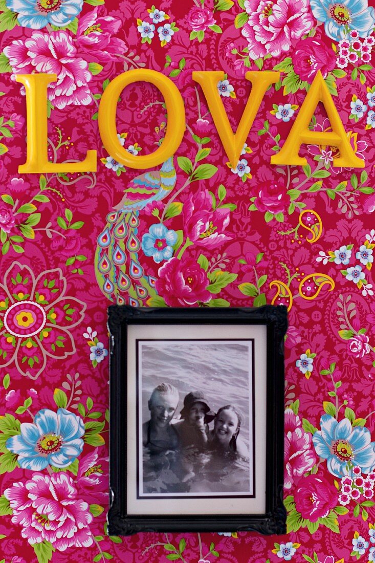 Gerahmtes Familienfoto unter gelben Deko Buchstaben auf tapezierter Wand mit Blumenmuster auf pinkfarbenem Hintergrund