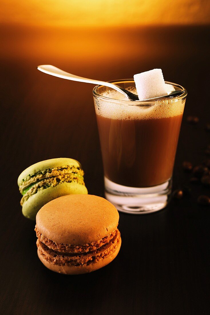 Kaffeeglas mit Löffel und Zuckerwürfel neben zwei Macarons