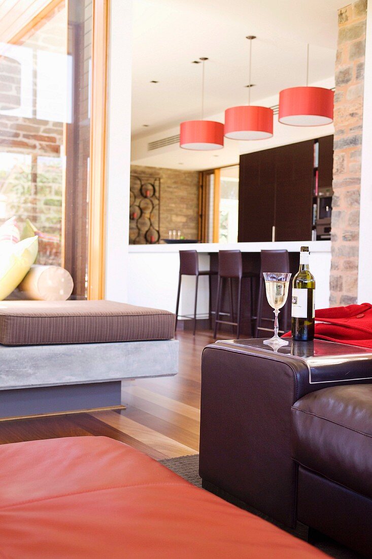 Detail of lounge area with leather-covered seating; open-plan kitchen in background with counter-style dining table below red, pendant lamps