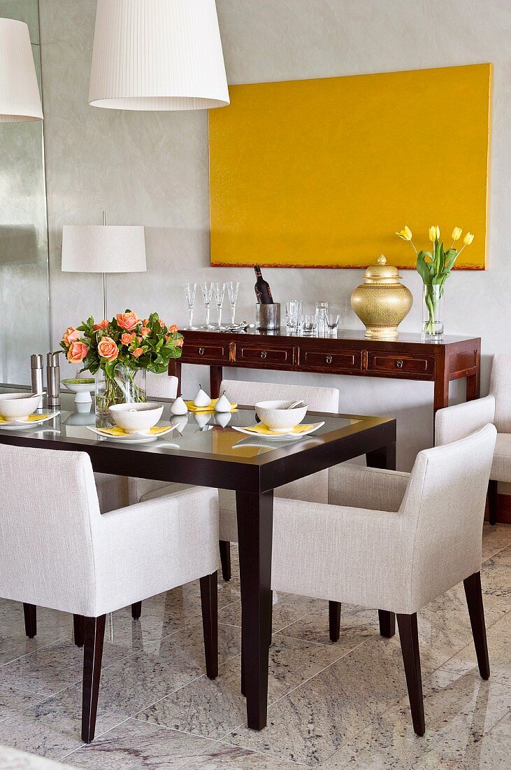 Ecru upholstered chairs around set dining table in front of antique sideboard below monochrome yellow artwork on wall