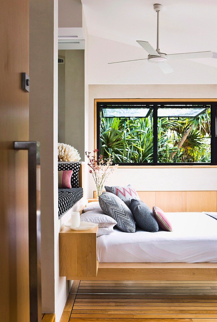 View through open door of stacked pillows on floating modern bed and palm trees seen through open window