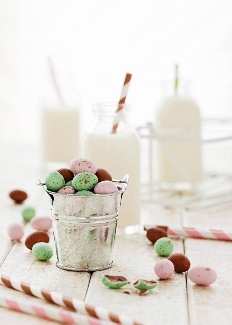 A mini bucket of pastel coloured chocolate eggs with small bottles of milk in the background