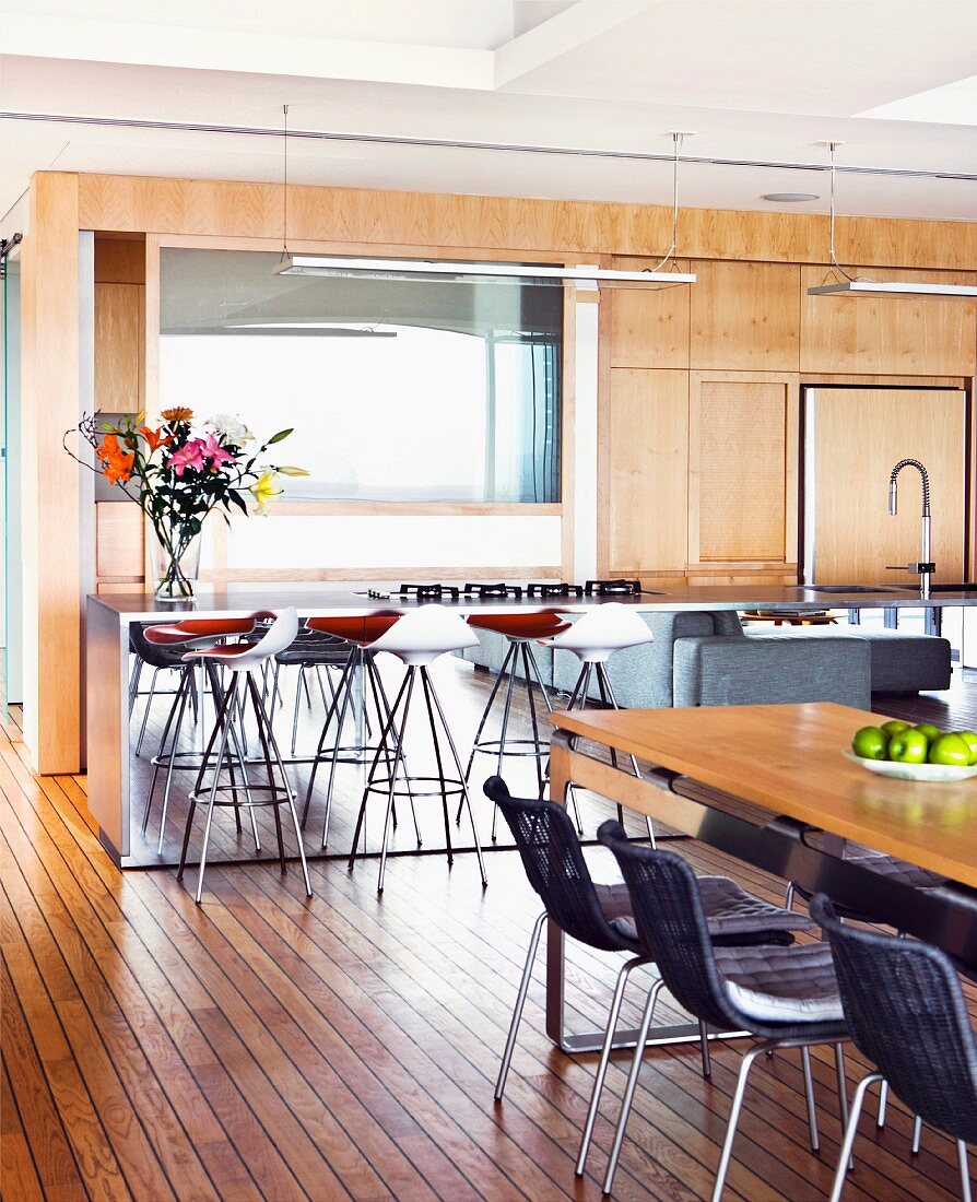 Dining table, dark wicker chairs and designer bar stools at kitchen counter in front of wooden, flat-fronted fitted kitchen cabinets