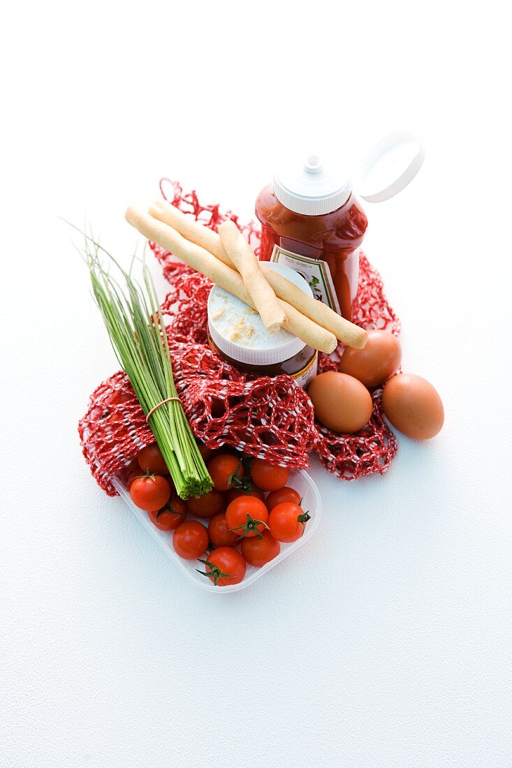A net shopping bag with tomatoes, chives, bread sticks, eggs and sauces