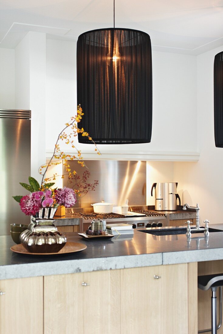 Pendant lamp with black, fabric lampshade above counter with sink and vase of flowers; illuminated gas stove in background