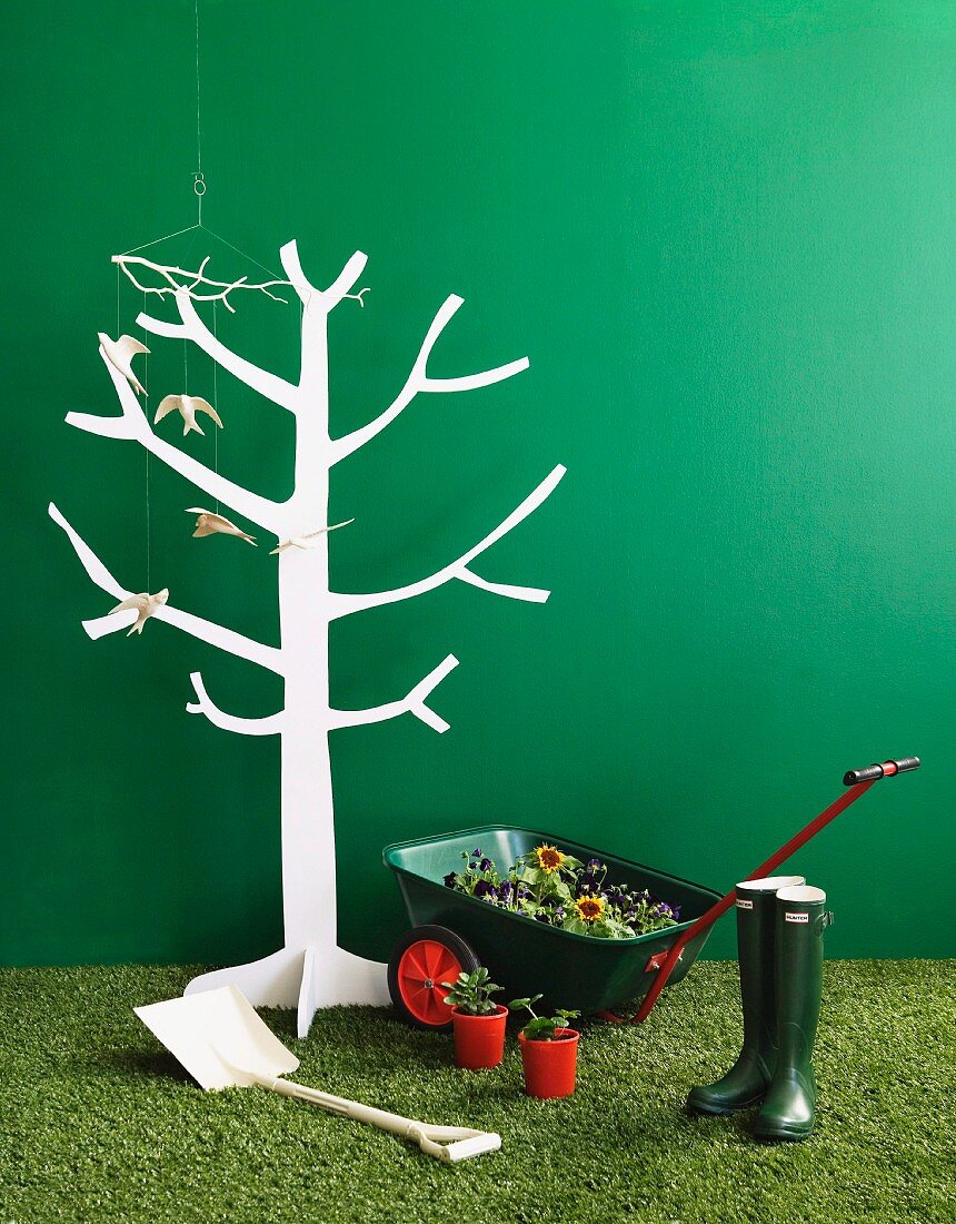 White, stylised wooden tree, flowers in wheelbarrow and gardening utensils on artificial lawn against green wall