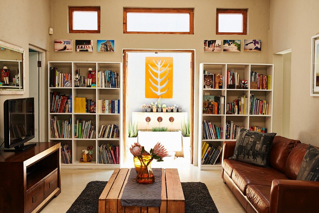 Living room with leather couch and wooden table; view of decorative, yellow artwork through open terrace door flanked by bookcases