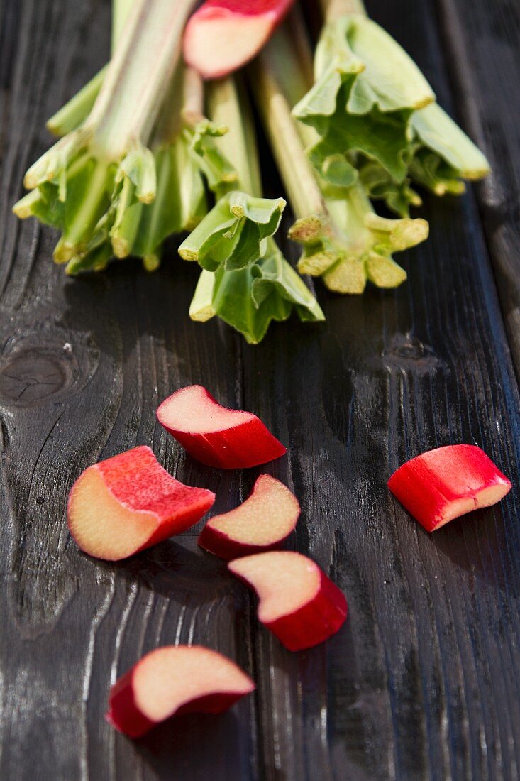 Pieces of rhubarb on a wooden surface with rhubarb stalks in the background