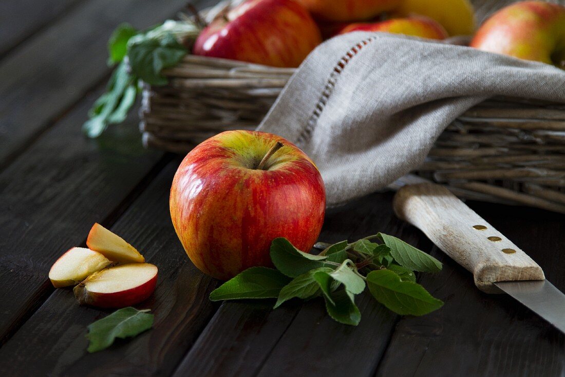 An apple with leaves in front of an apple basket with a few pieces of apple next to it