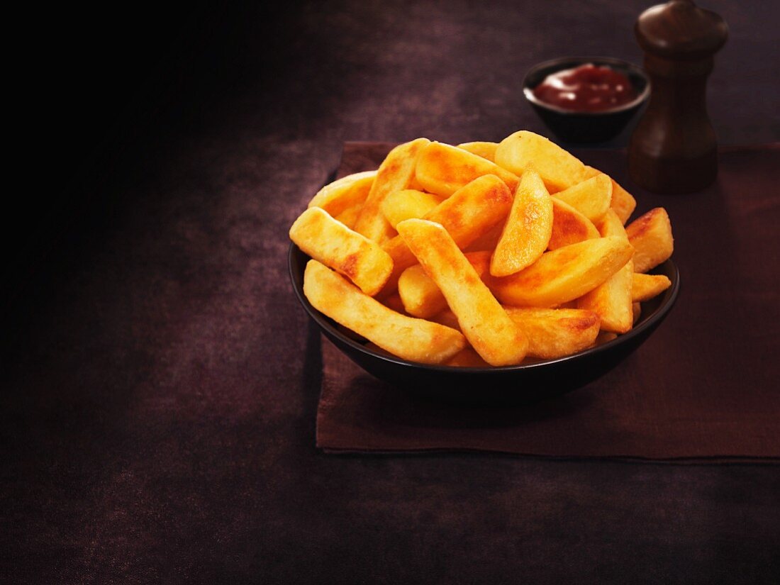 A bowl of thick-cut chips with ketchup and pepper