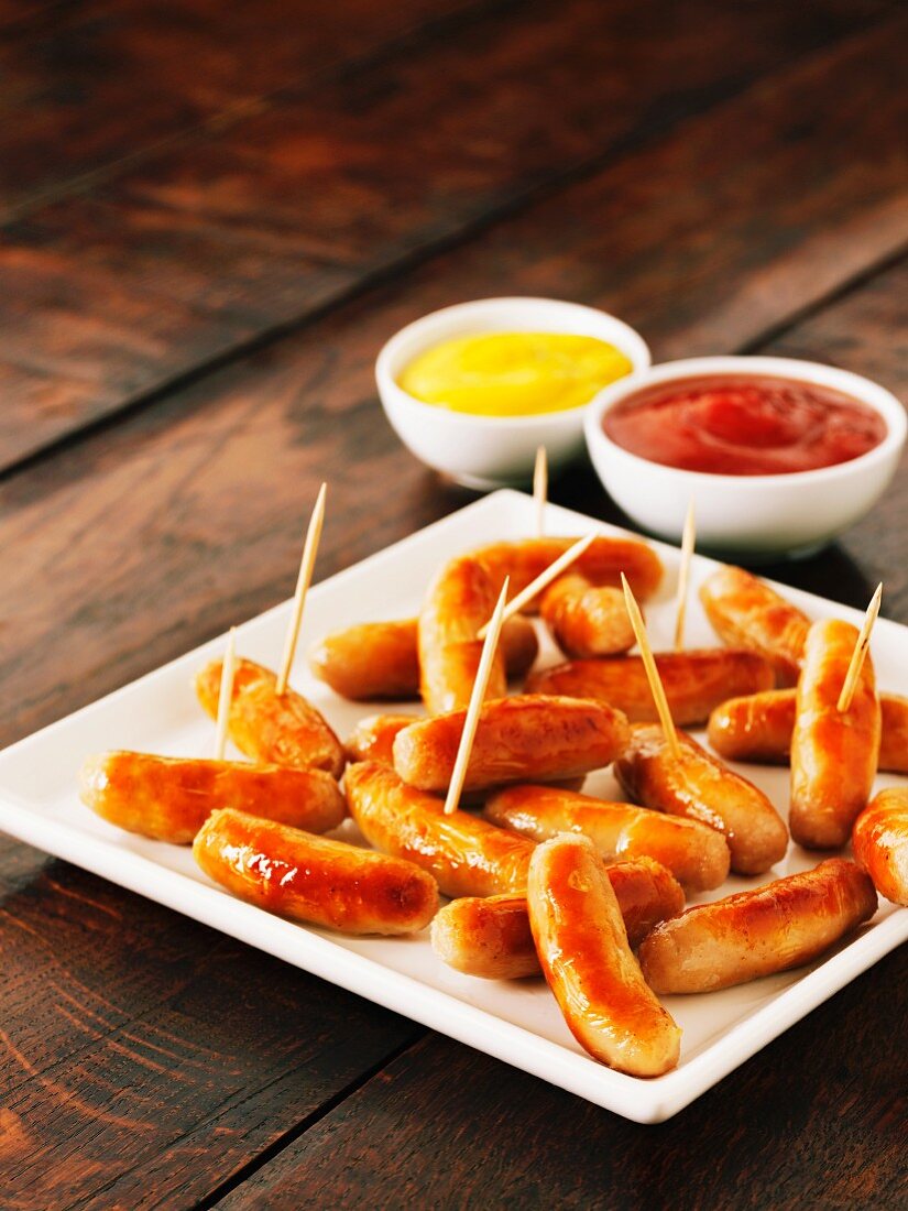 Fried cocktail sausages on sticks on a plate with dishes of ketchup and mustard