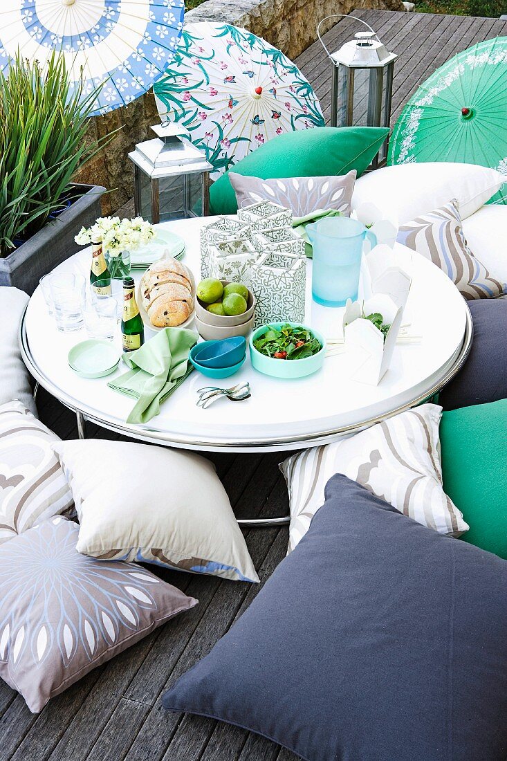 Food and drinks on round, low table surrounded by various floor cushions on wooden deck