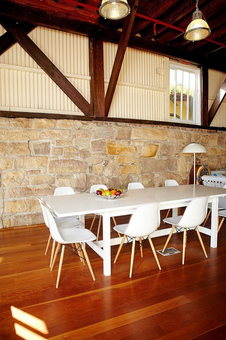 A dining area with white bucket chairs in a hall-like living room with a half-height natural stone wall and visible roof beams