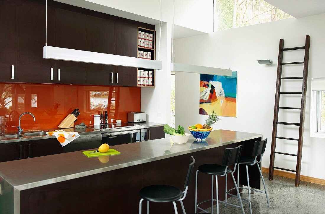 Dark brown designer kitchen with orange glass splashback, stainless steel worksurface on counter with bar stools