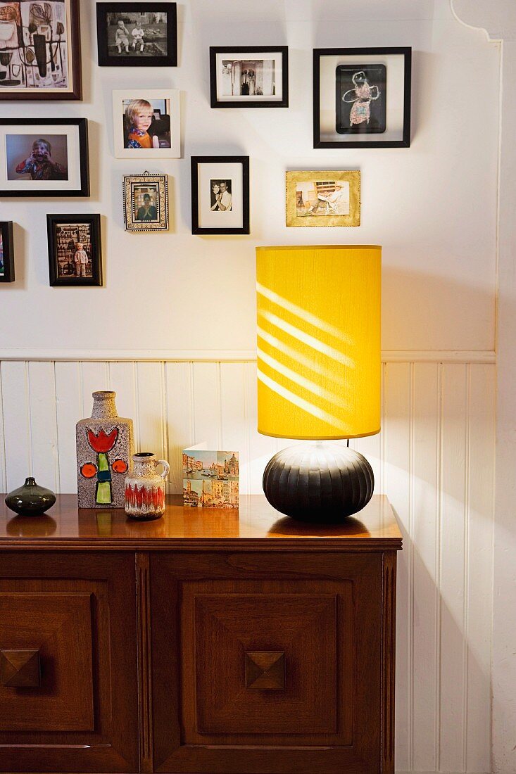 Table lamp with yellow fabric lampshade on wooden sideboard against white wainscoting and below framed photos