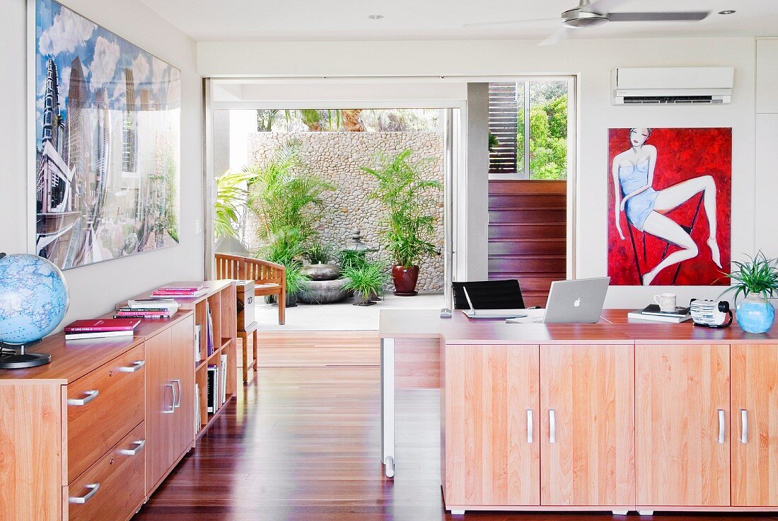 Open-plan office with sideboards, artworks on walls, open terrace doors and staircase in background