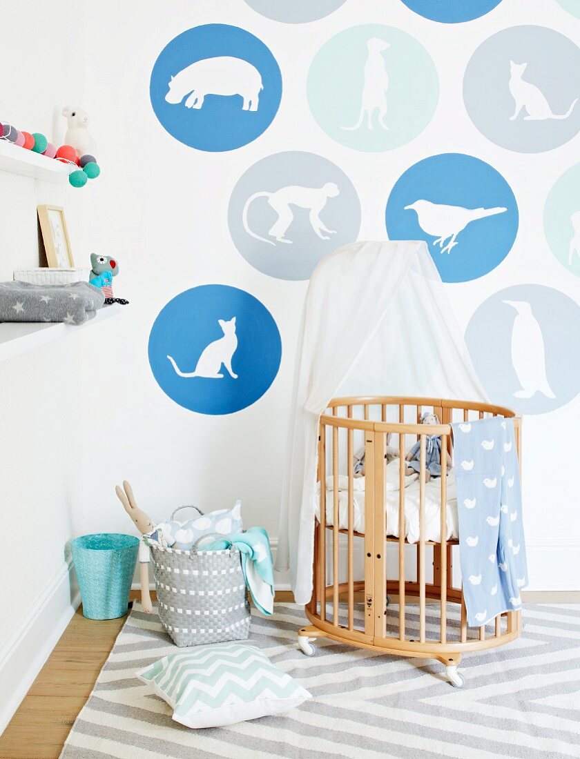 Designer cot made from pale wooden rods with canopy against wall with animals motifs in circles of colour