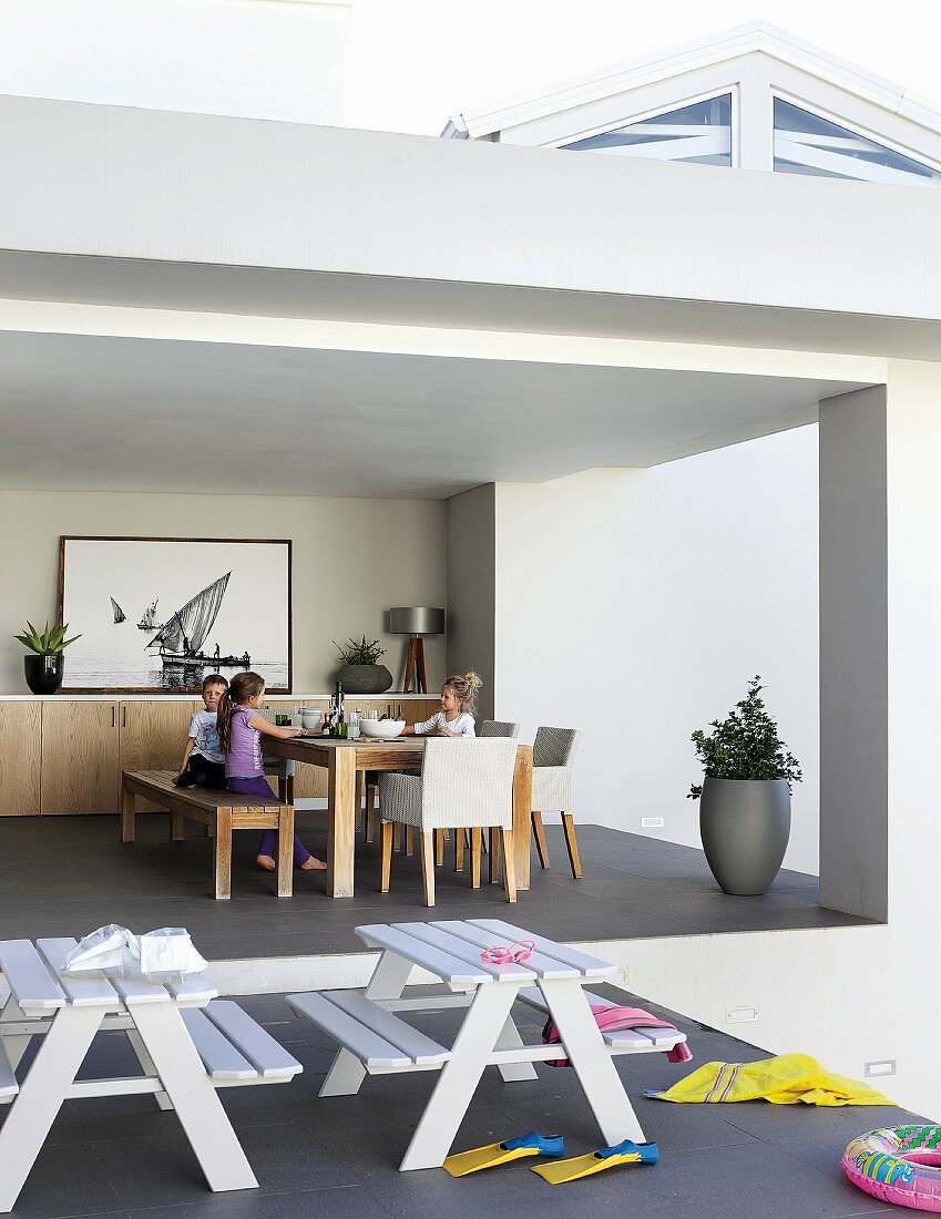 Children in dining area and sideboard on comfortable, roofed terrace; two children's picnic bench combinations and scattered swimming gear in foreground