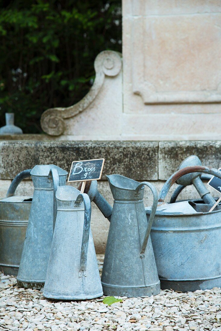 Old zinc jugs with price labels on a gravel surface in front of an antique wall