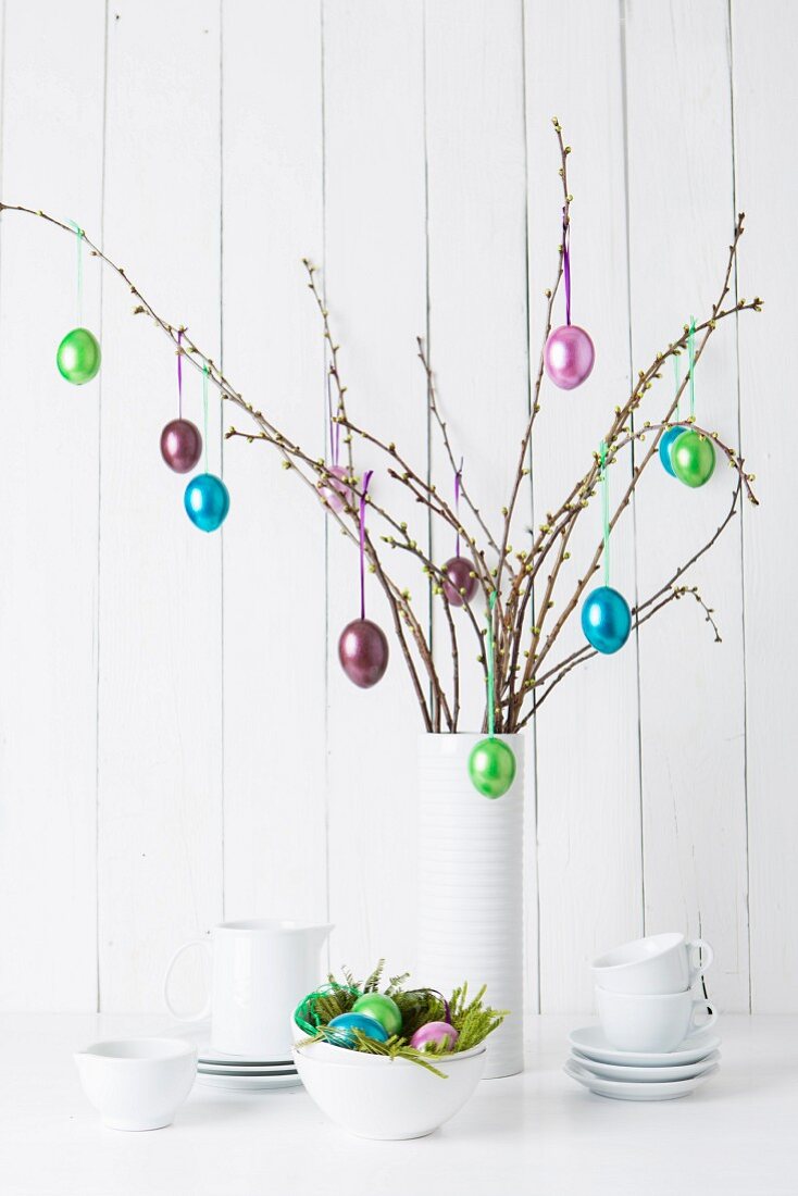 Still-life arrangement of crockery and glossy Easter eggs hanging from twigs