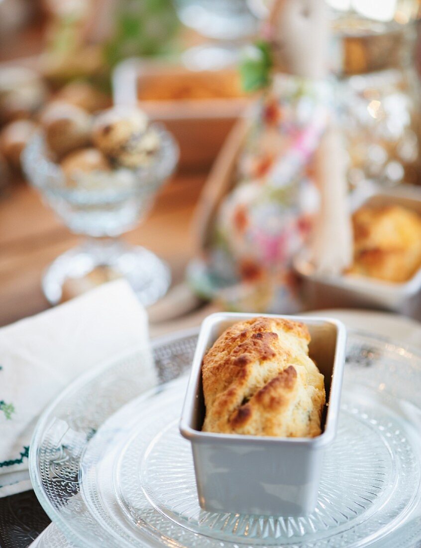 A mini loaf of cheese and thyme bread in a baking tin on a table laid for Easter