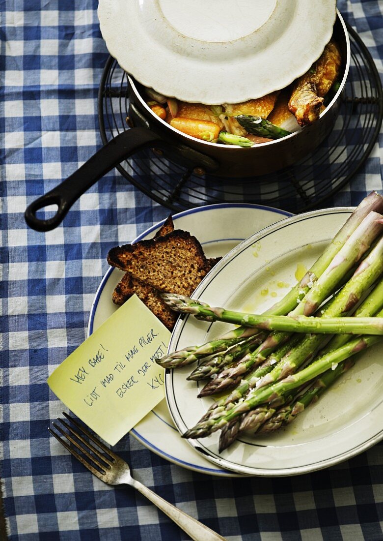 Green asparagus and grilled bread on plates with a sticky note and a saucepan of chicken in the background