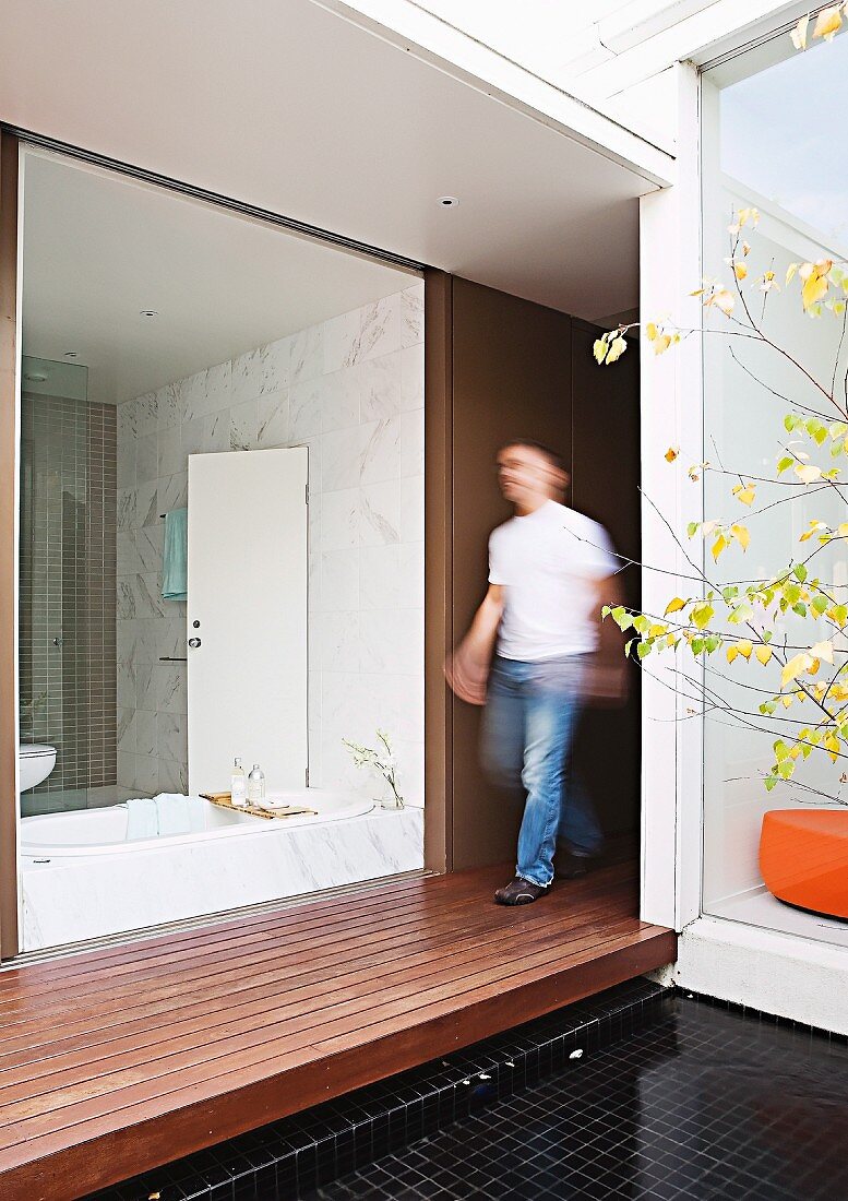 View from courtyard with wooden platform through frameless glass wall into white bathroom opposite home office; blurred man walking between the rooms