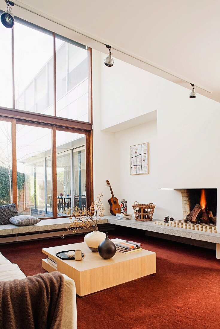 Seating area in front of fireplace with cubic wooden table on rust red carpet, encircling concrete bench and sofa in room with double-height glass facade