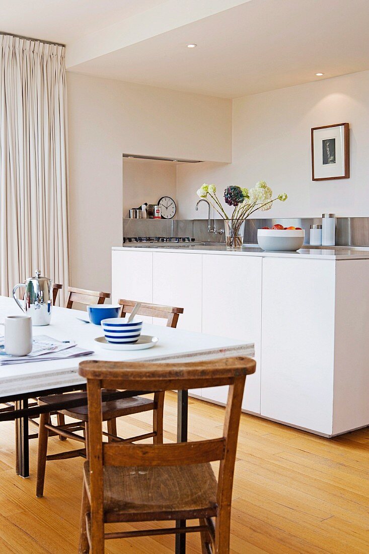 Table set for breakfast and simple wooden chairs in front of island counter in purist, white, designer kitchen